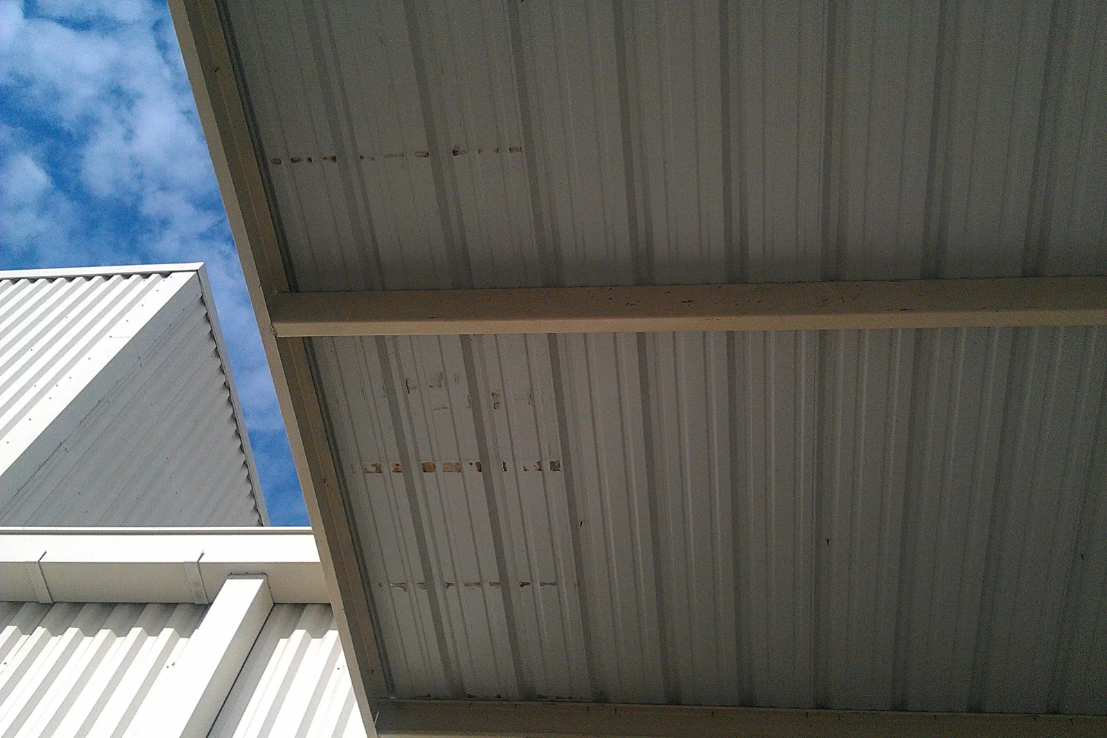 Underside of white corrugated metal carport roof with painted steel beam and ribbed panels against blue sky