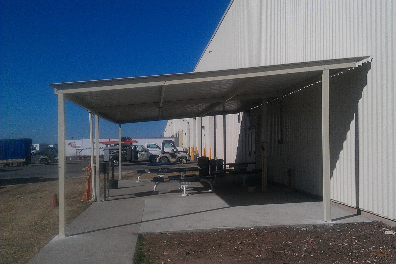 Cream steel lean-to canopy with corrugated metal roof over concrete slab beside industrial metal building wall