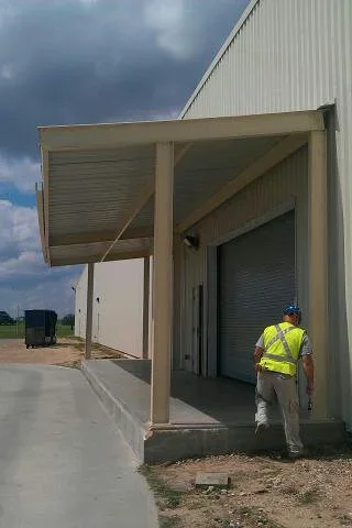Tan metal canopy with corrugated roof over roll-up door on steel building with concrete pad