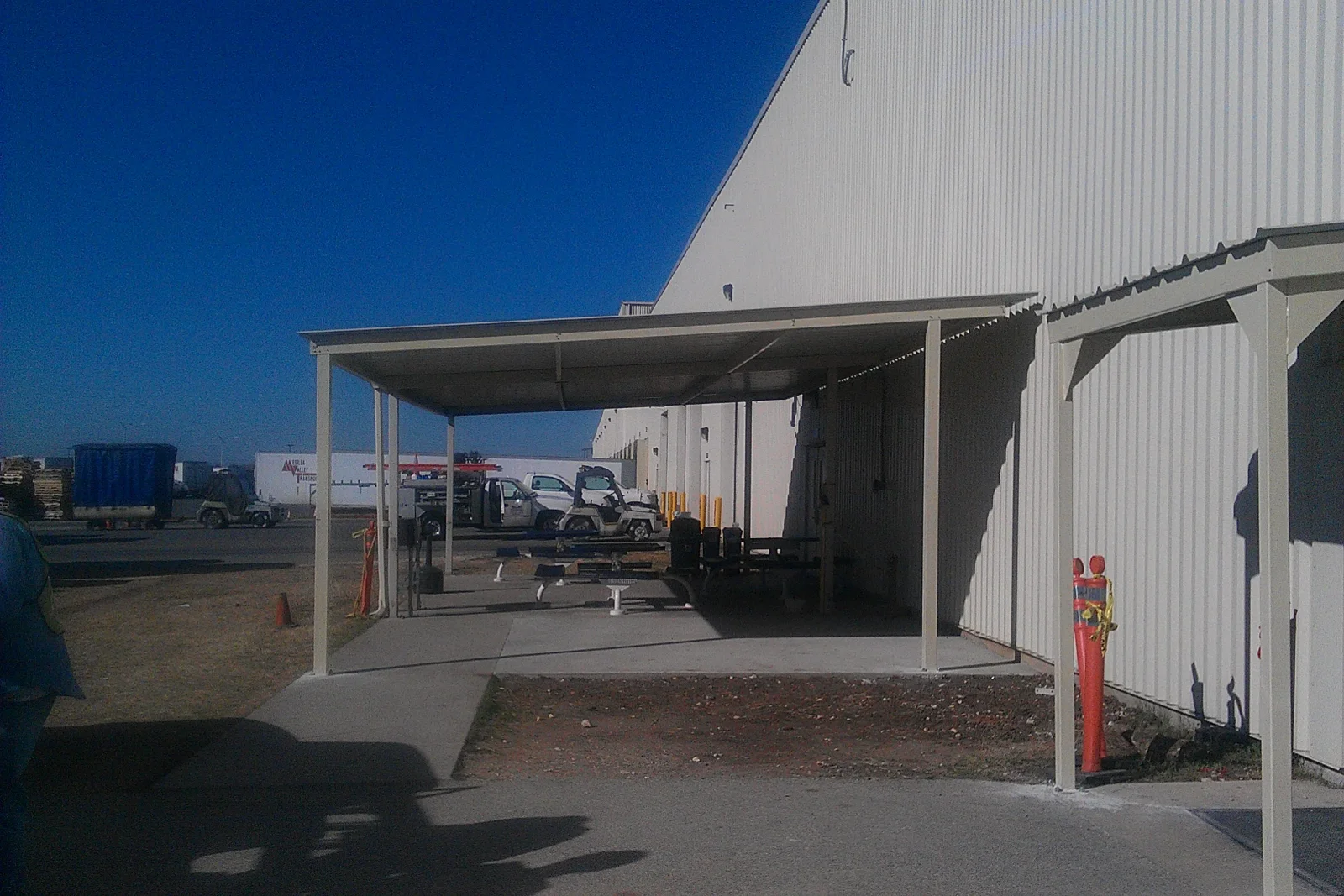 Beige steel cantilever canopy with square columns and metal roof panels attached to a corrugated metal building over a concrete pad
