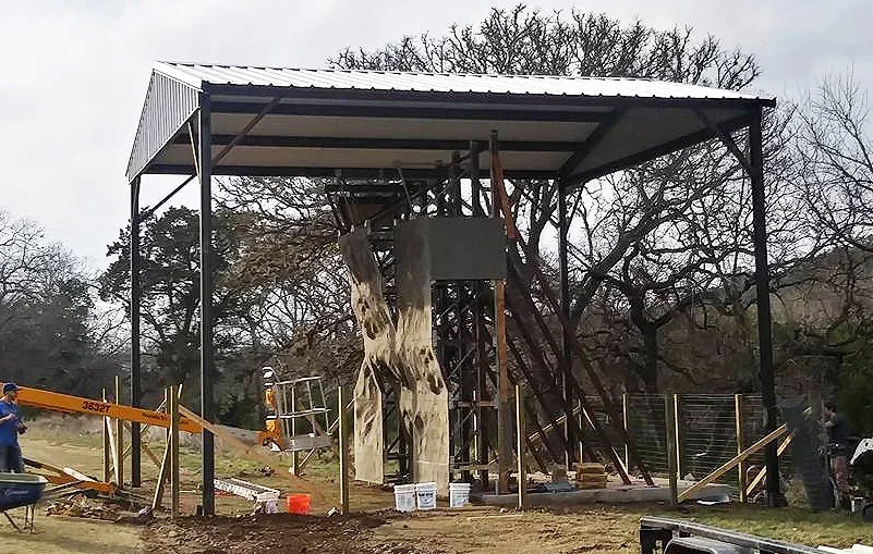 Black steel carport frame with galvanized metal roofing under construction beside a textured tan column and wooden stakes