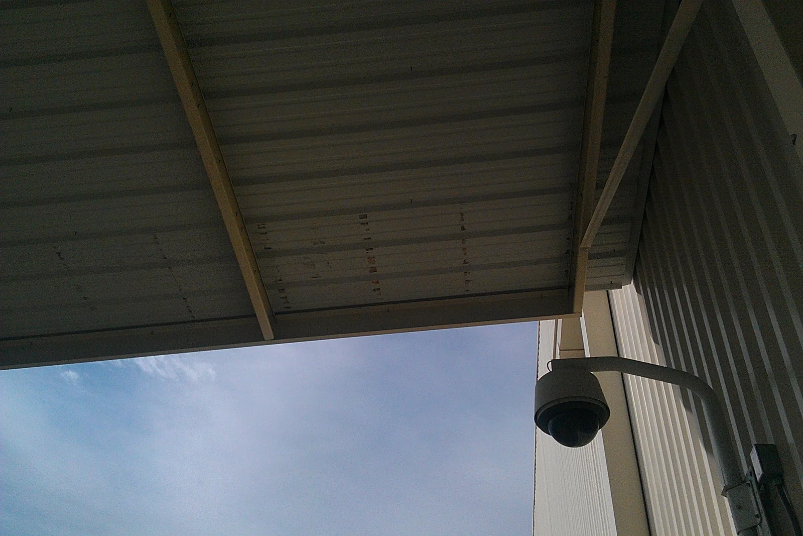 Underside of corrugated metal canopy panels on exposed wood beams with a dome camera mounted on a bracket