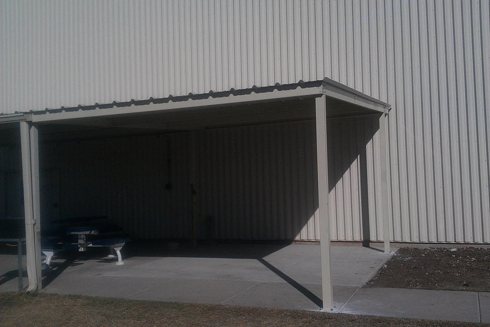 Light-colored steel lean-to carport with dark ribbed metal roof panels over a concrete slab beside a corrugated metal building