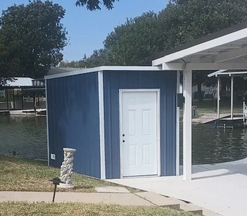 Blue vertical-siding storage building with white trim and door beside a white carport on a concrete slab