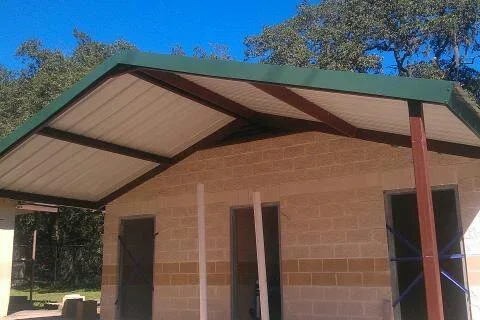 Gabled carport with cream corrugated roof, green metal trim, and tan split-block storage room with dark doors