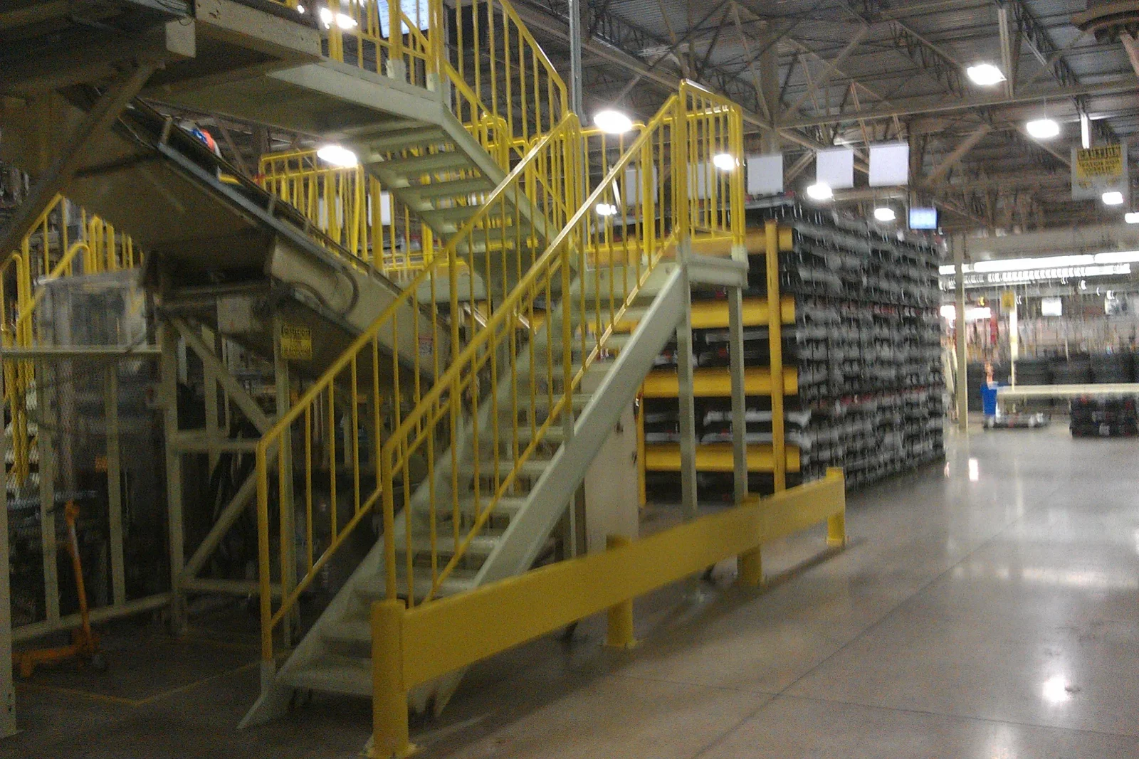 Steel staircase with yellow guardrails and grey treads beside yellow pallet racking in a warehouse