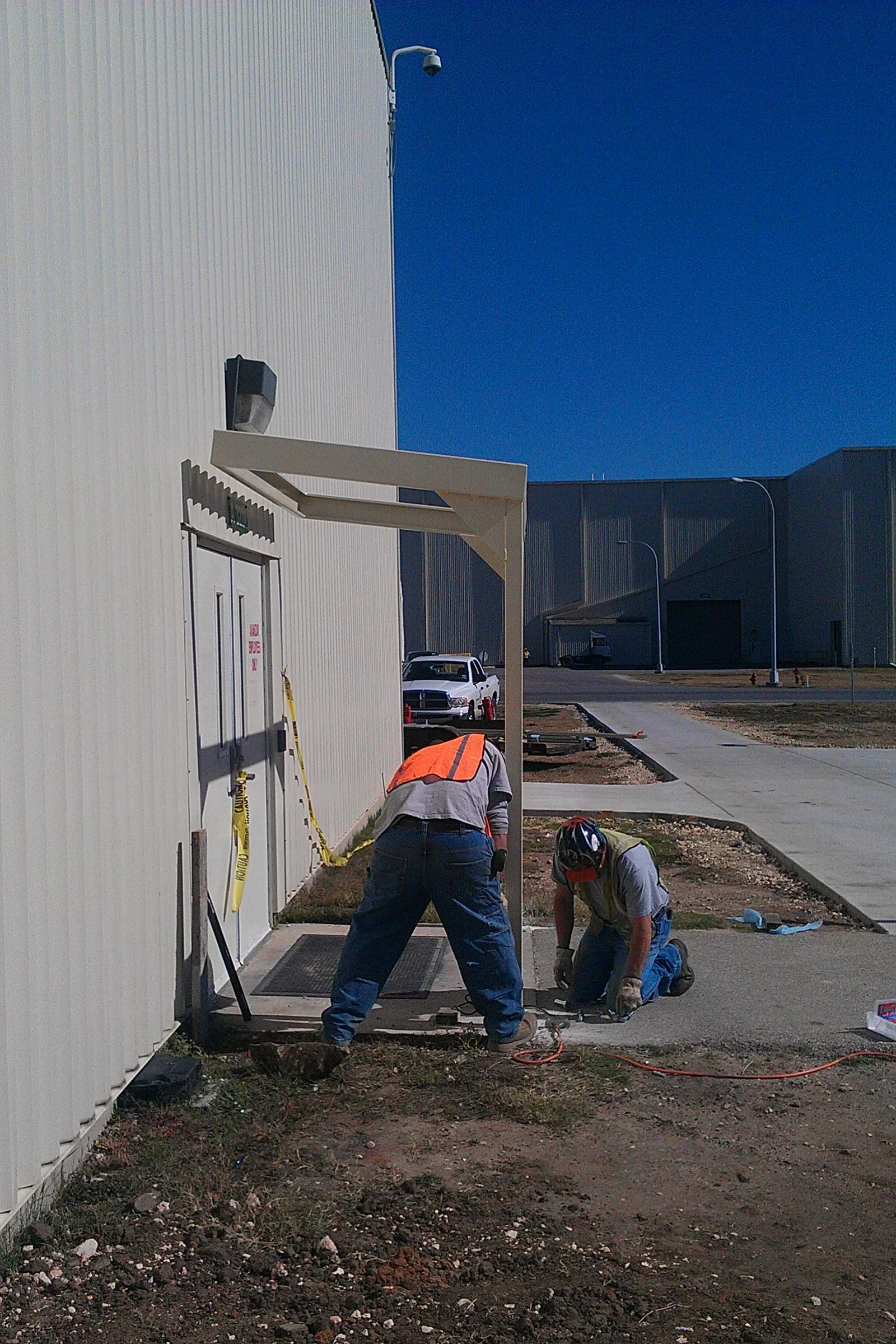 Workers installing a cream steel entry canopy over a metal door on a corrugated metal building with concrete pad