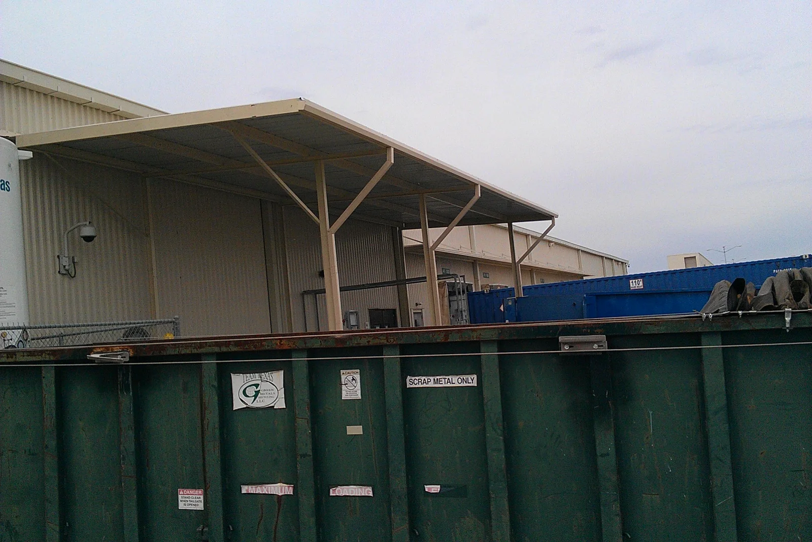 Cream painted steel canopy with sloped metal roof and diagonal braced posts over an outdoor loading area beside a corrugated metal building