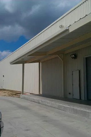 Tan corrugated metal commercial building with covered canopy, timber posts, concrete loading dock, and roll-up door