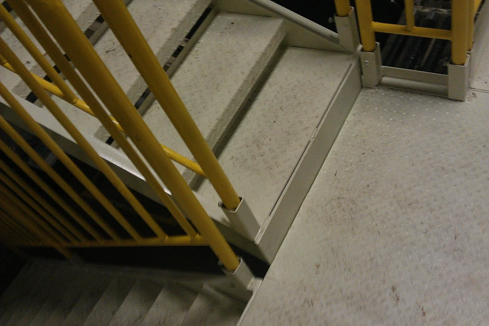 Gray steel staircase with diamond-plate treads and yellow painted tubular guardrails on a metal landing