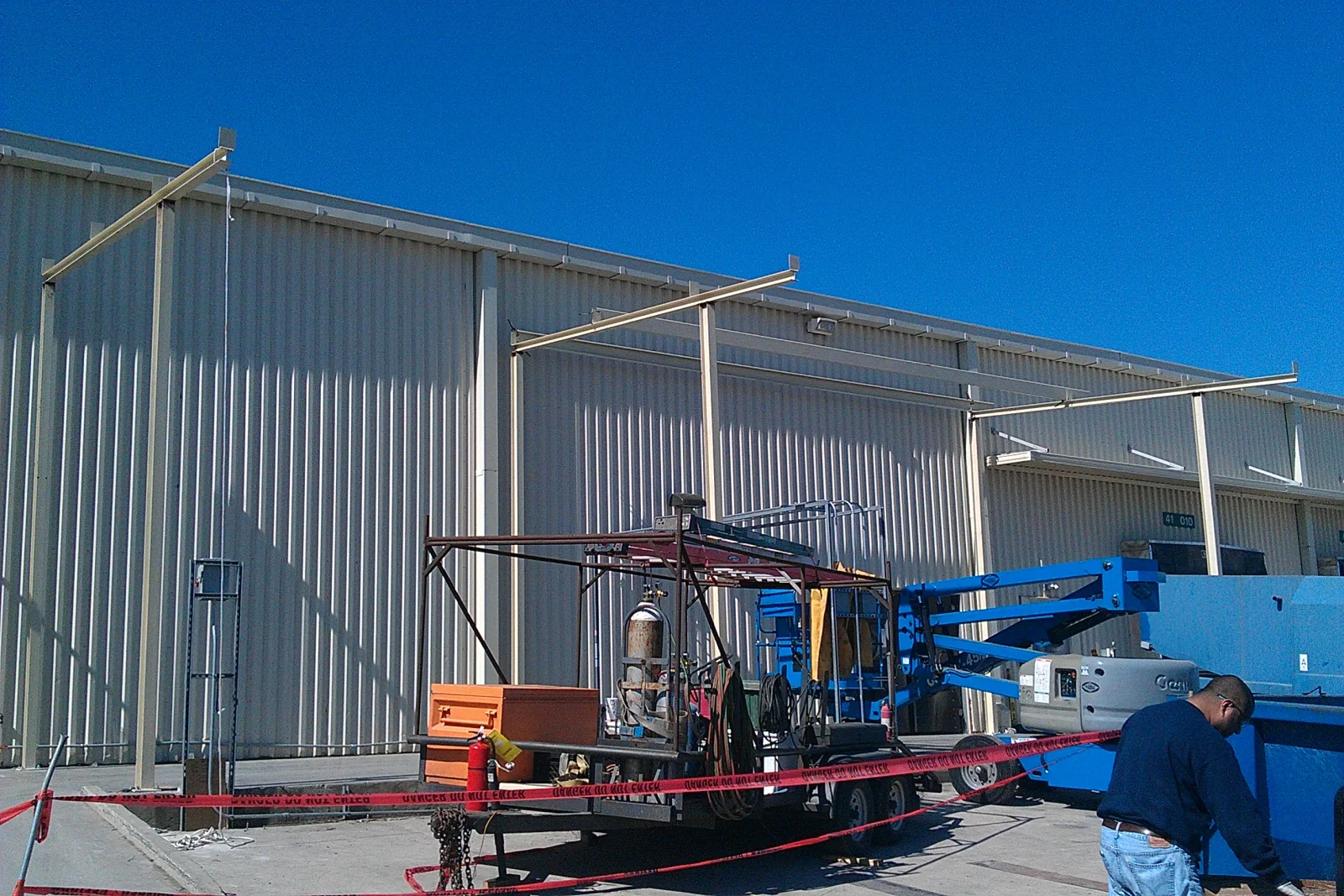 Steel columns and cantilever beams being installed for a canopy against corrugated metal building, with boom lift and welding trailer on site