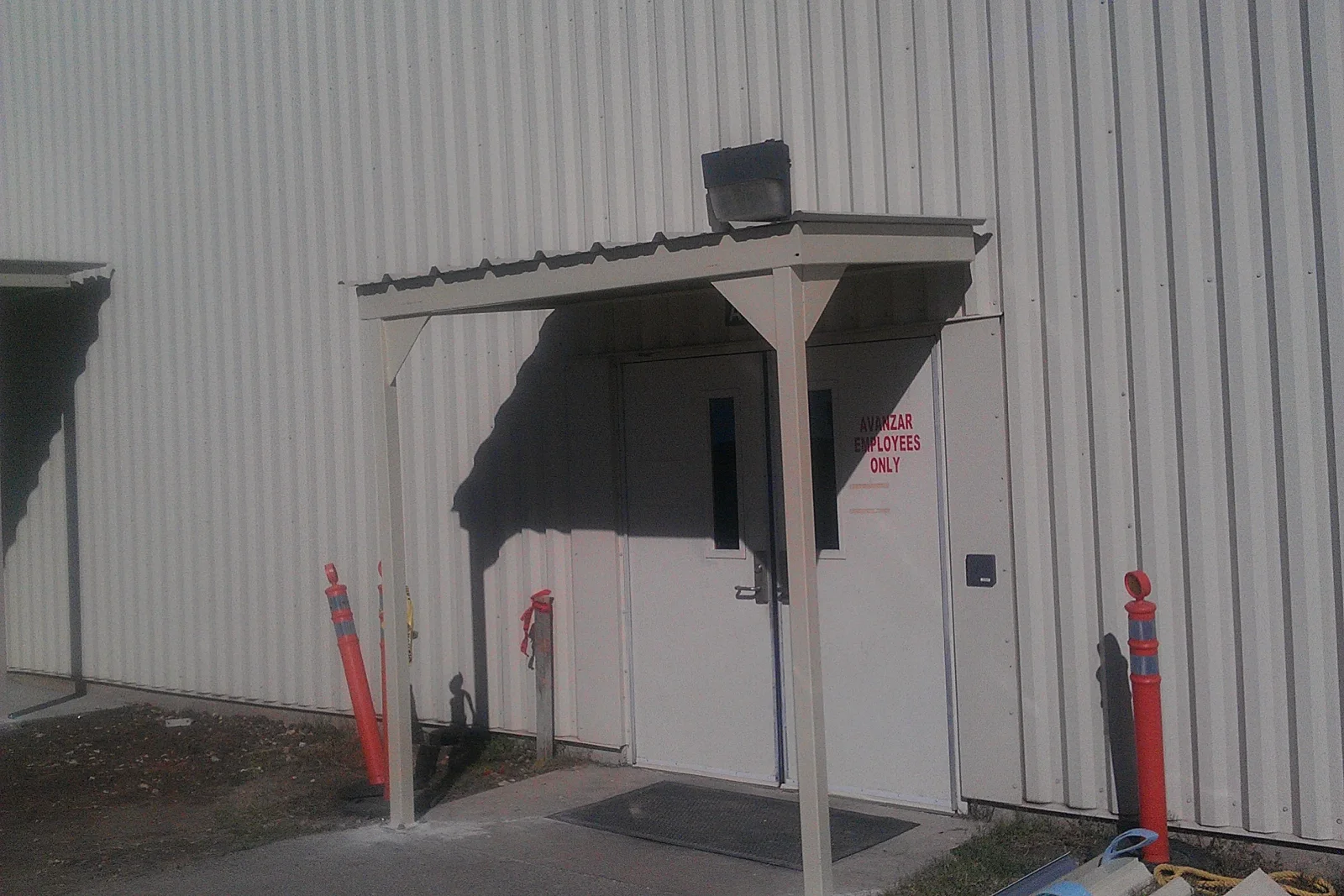 Beige steel entry canopy with square posts and metal roof over a steel door on corrugated metal wall