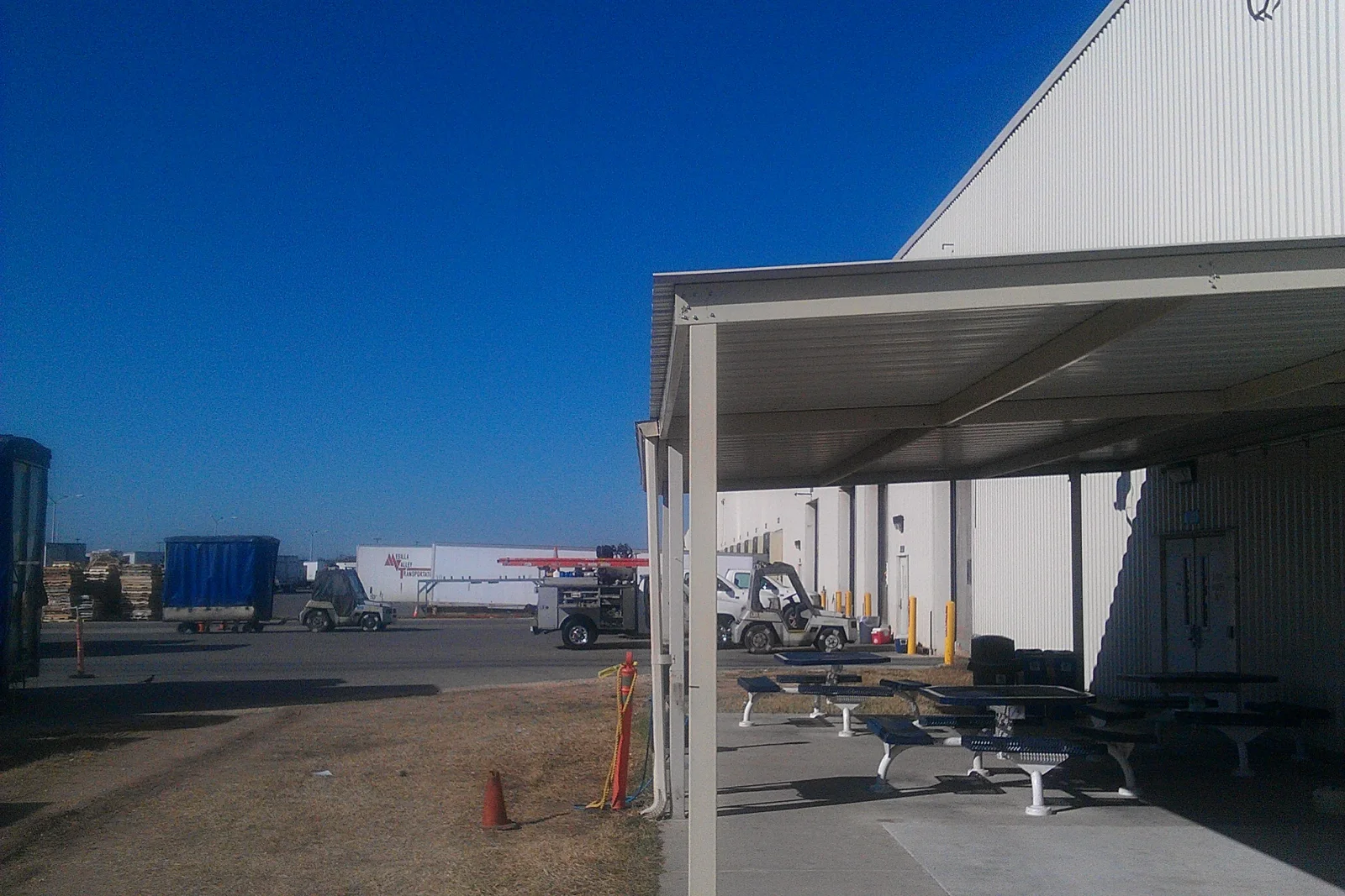 Steel canopy over concrete patio with metal picnic tables beside corrugated metal building