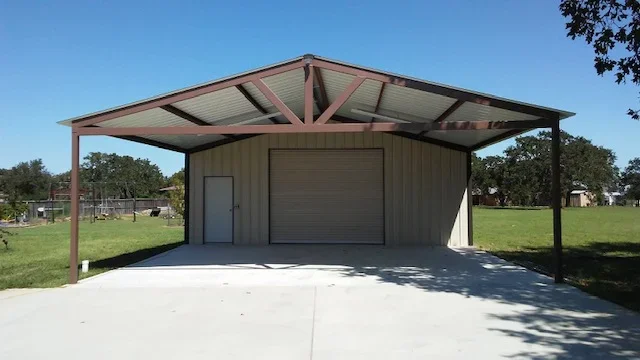 Tan steel-panel garage with roll-up door and brown metal carport roof supported by steel posts over concrete pad