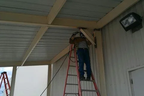Worker on stepladder attaching tan beam to underside of corrugated metal carport roof with white metal wall panels