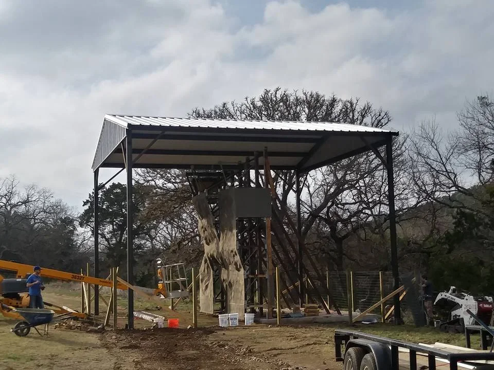 Dark steel carport frame with white metal roof panels under construction, with boom lift and skid steer on site