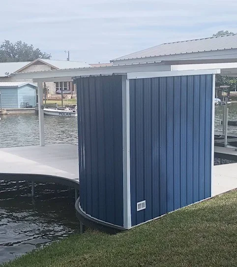 Blue corrugated metal storage shed with white trim on a curved concrete dock beside water, with a metal roof
