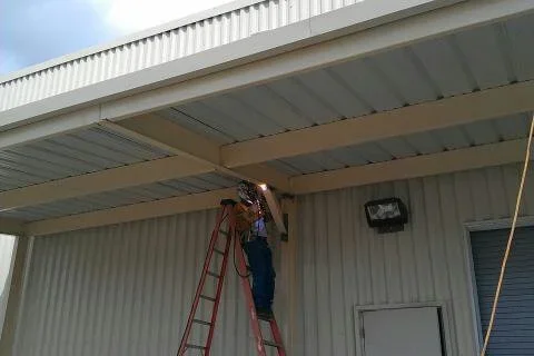 Worker on red ladder fastening white corrugated metal roof panels to beige steel carport framing